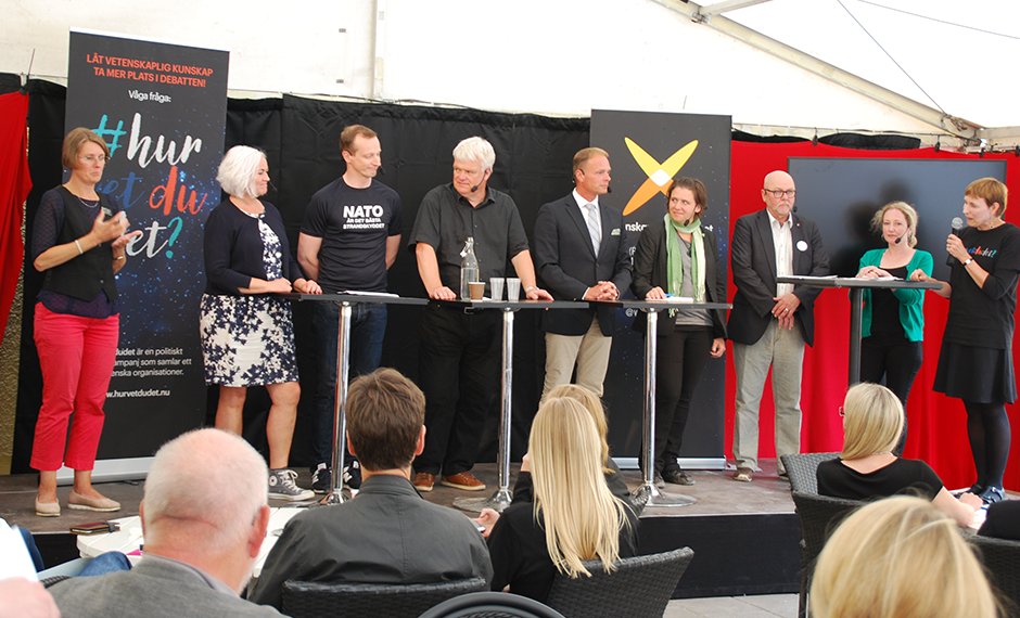Sju politiker står på en scen, med en teckentolk till vänster och moderatorn Cissi Askwall till höger.